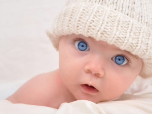 A cute little baby is looking into the camera and is wearing a white hat. The baby could be a boy or girl and has blue eyes. use it for a parenting or love concept.; Shutterstock ID 62715262; PO: novembre