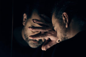 Man in despair, sad and lonely resting his head on a hand, in front of mirror